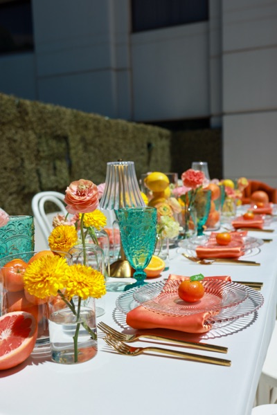An outdoor table in coral and turquoise, with scalloped glass plates, gold flatware, marigolds and sliced citrus.