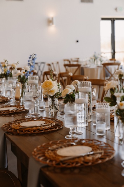 A long farmhouse table set with woven rattan chargers, ribbed glassware and low vases of butter-yellow roses and blue delphinium.